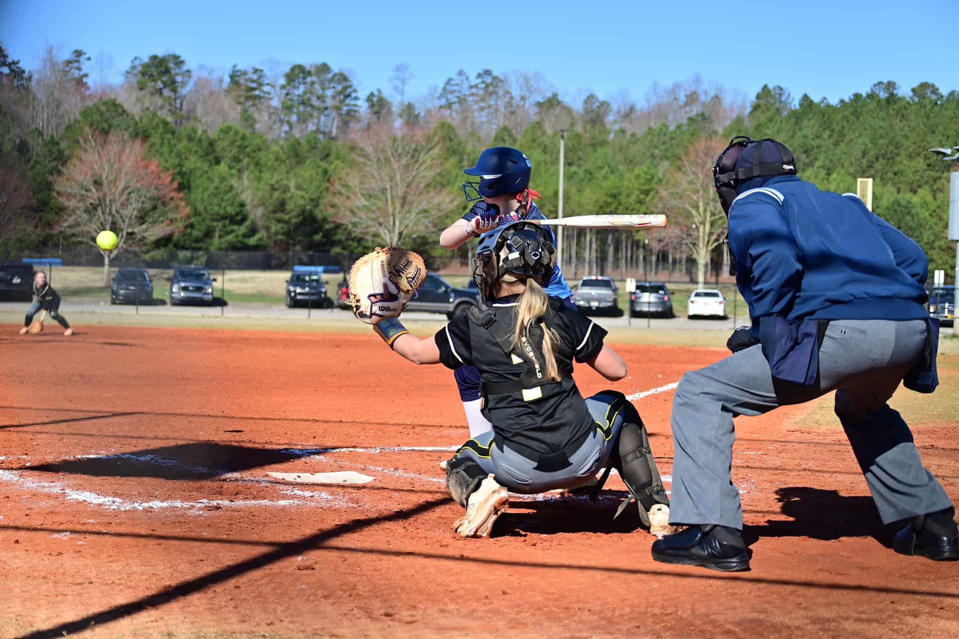 A softball catcher in full gear behind the plate as the batter waits on a sunny day